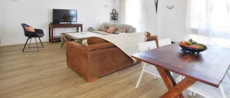 A modern living room with a wooden floor, a brown sofa, a white dining table, and a television on a stand.