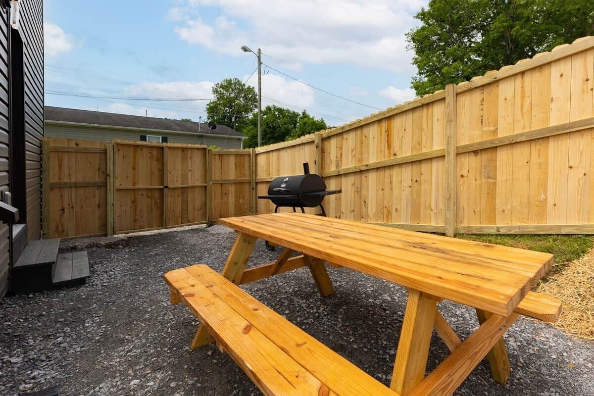 A wooden picnic table with benches in a backyard with a wooden fence and a barbecue grill.