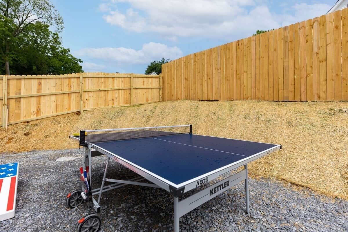 A ping pong table with wheels, labeled 'AXOS' and 'KETTLER', set up outdoors with a wooden fence and gravel ground.