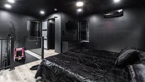 Une chambre avec un couvre-lit en cuir noir, un ventilateur de plafond et une petite borne d’arcade rose.