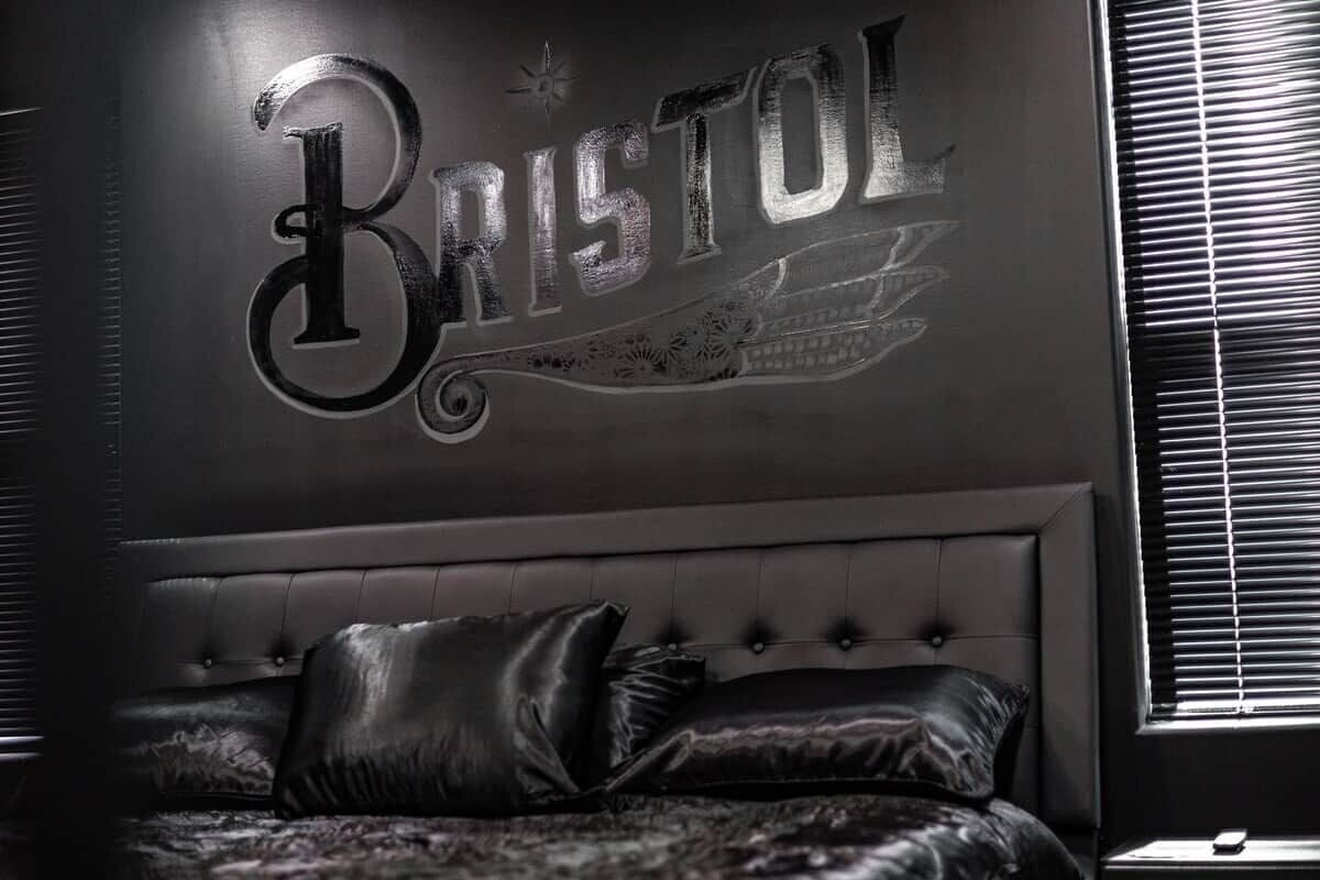 Een slaapkamer met een groot bed, een bord aan de muur met de tekst 'BRISTOL' en een raam met jaloezieën.