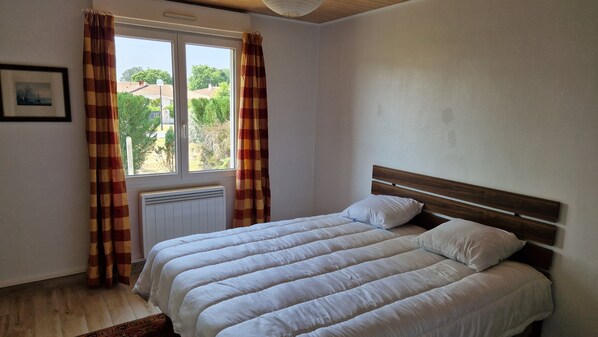 A bedroom with a bed, curtains, a radiator, and a window with a view of greenery. - La Villa du Marensin (Lit-et-Mixe)