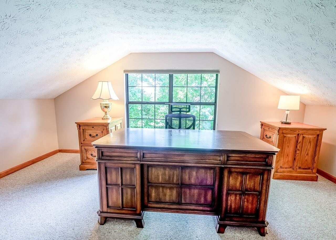A room with a large wooden desk, two wooden nightstands, a window with a view of greenery, and a ceiling with a textured pattern.