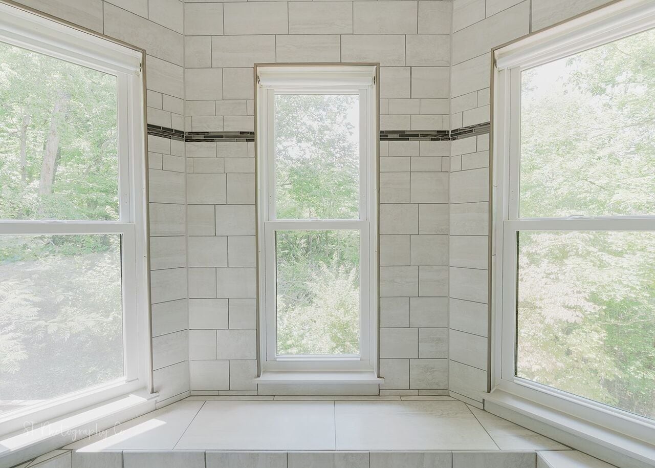 A corner room with three large windows, white tiled walls, and a visible tree outside.