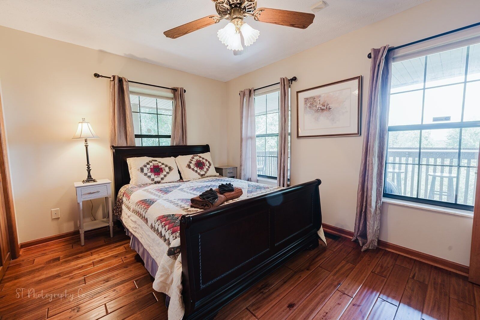 Une chambre avec un plancher en bois, un lit recouvert d’une courtepointe, une table de chevet avec une lampe, un ventilateur de plafond et une fenêtre avec des rideaux.