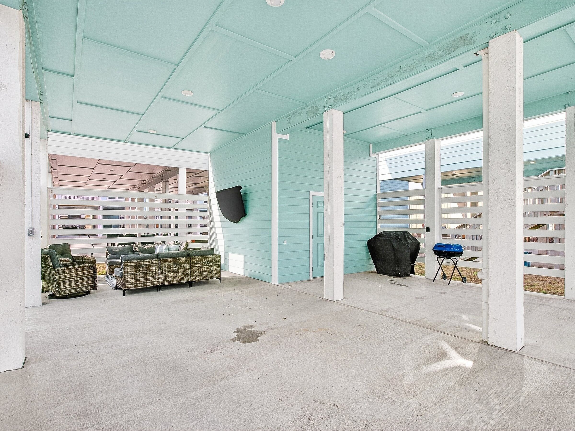 A covered outdoor area with a concrete floor, a wicker sofa, a grill, and a white fence.