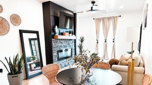 Un salon moderne avec une table ronde, des chaises en osier, un canapé et un téléviseur fixé sur un mur noir.