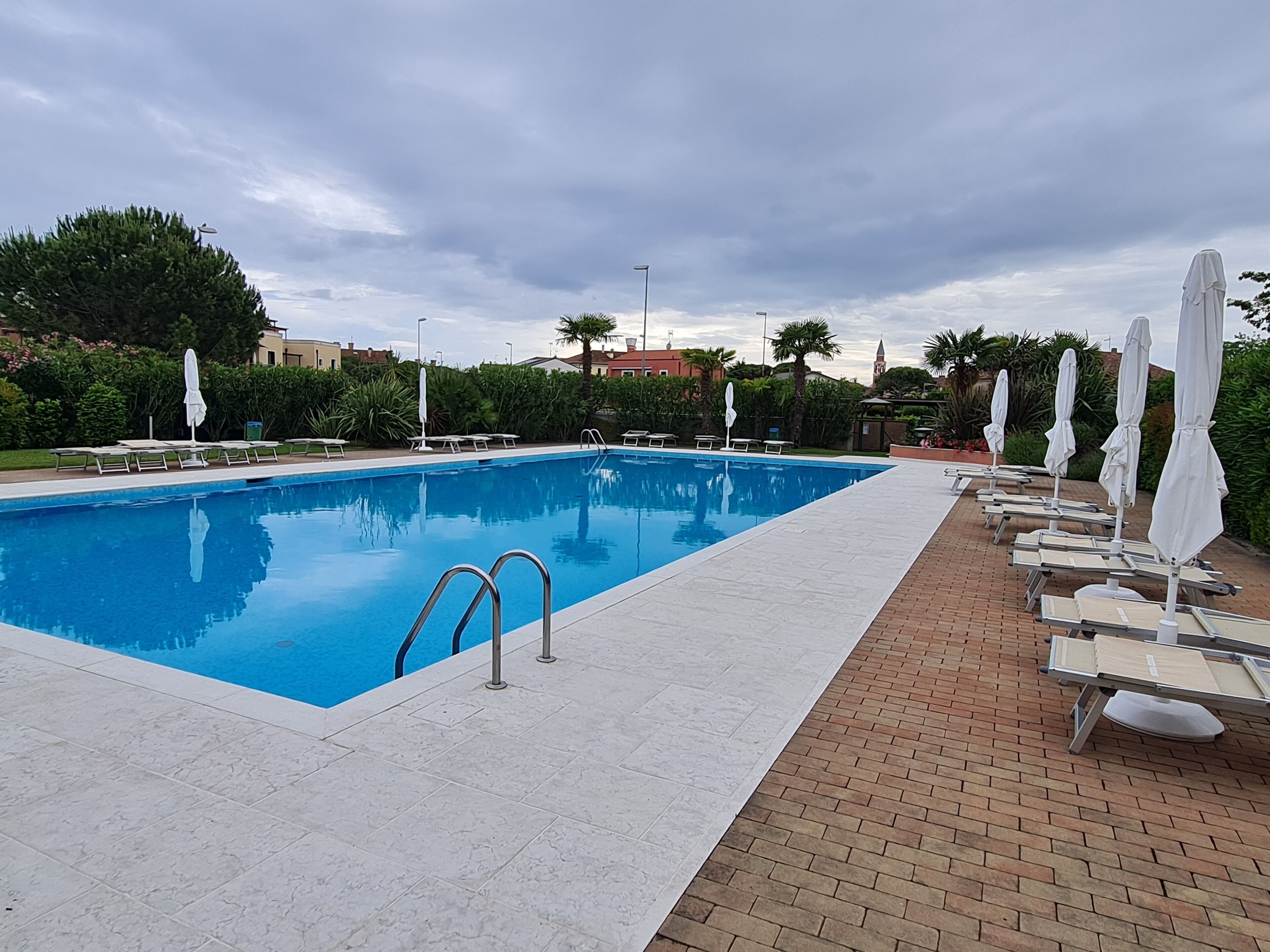 A rectangular swimming pool with clear blue water, surrounded by a tiled deck. There are lounge chairs with white umbrellas along the poolside.
