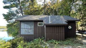 A small cabin with a window and a door, surrounded by trees and a body of water.