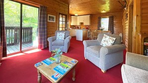 Een cabinte interieur met twee grijze fauteuils, een houten bijzettafeltje met tijdschriften, een keukenhoek en een schuifdeur van glas naar buiten.