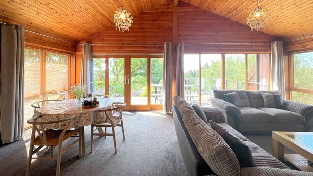 En hyggelig hytteinteriør med træloft, spiseplads med bord og stole, sofa og store vinduer der giver udsigt til naturen.