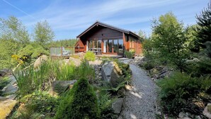 En træhytte med terrasse og en grusvej, omgivet af grønt og sten.