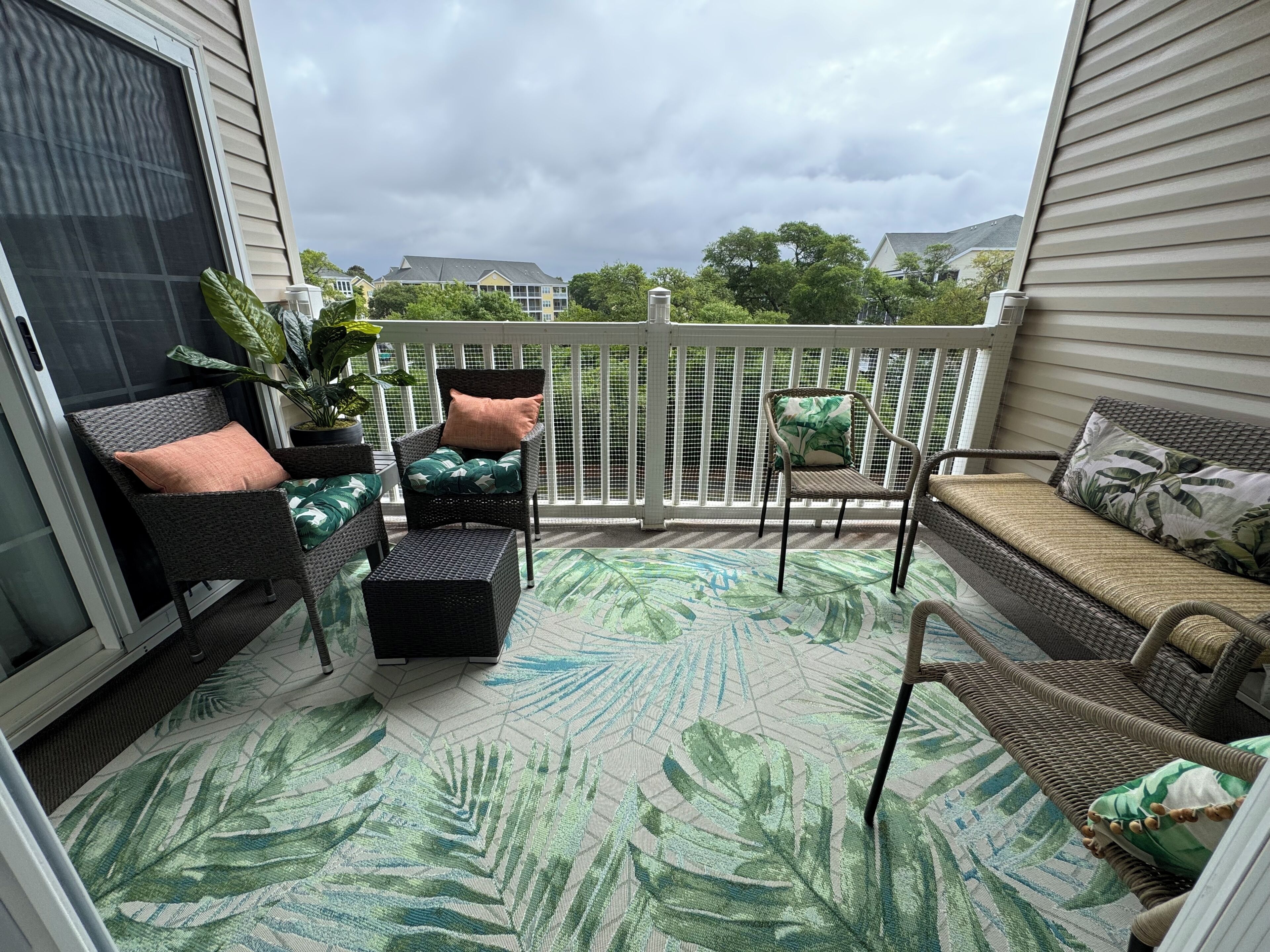 A covered outdoor seating area with wicker furniture, a patterned rug, and a white fence.