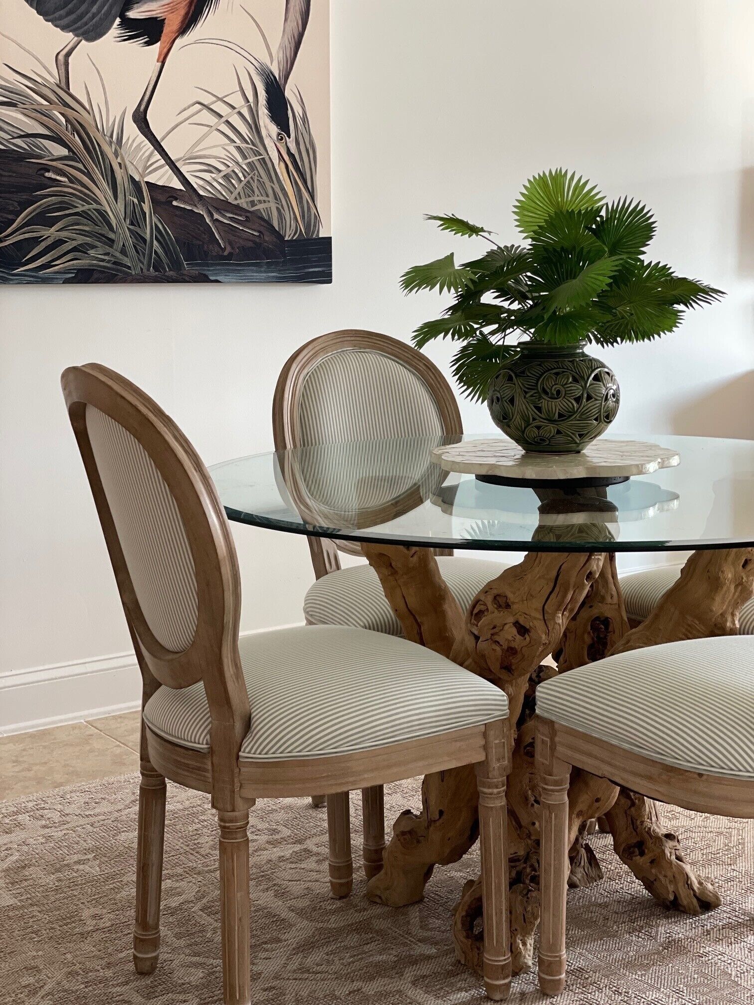 A dining table with a glass top surrounded by chairs with cushioned seats, a decorative vase with a plant, and a piece of wall art depicting a bird.