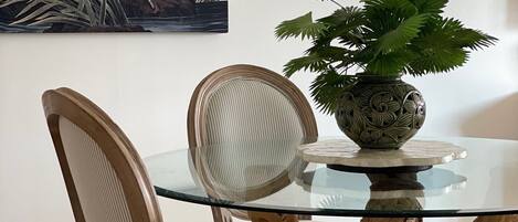 A dining table with a glass top surrounded by chairs with cushioned seats, a decorative vase with a plant, and a piece of wall art depicting a bird.