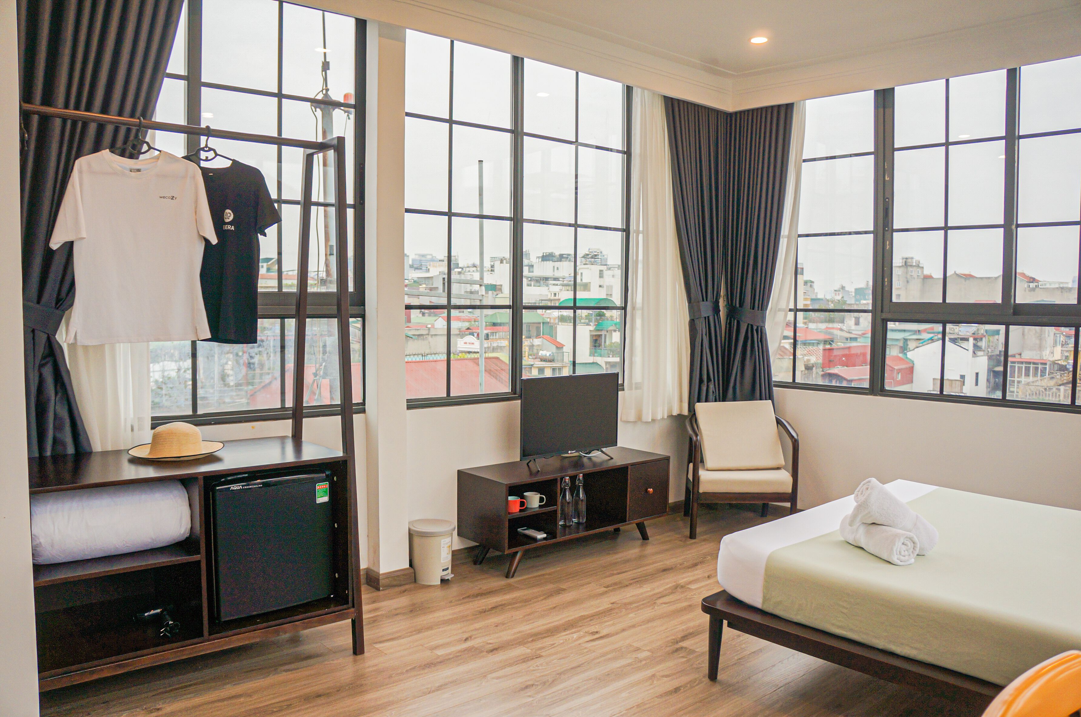 A hotel room with a bed, a TV, a small fridge, a chair, and a window with a city view.