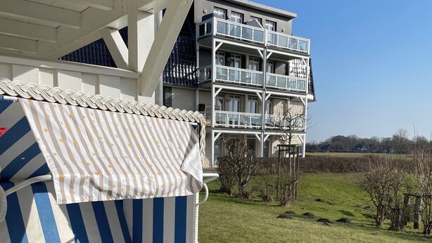 A covered outdoor seating area with a striped sofa and a table, overlooking a grassy field and a multi-story building. - Apartment "Moin im Deichgraf" on the Wremer beach and cutter harbor (Wurster Nordseeküste)