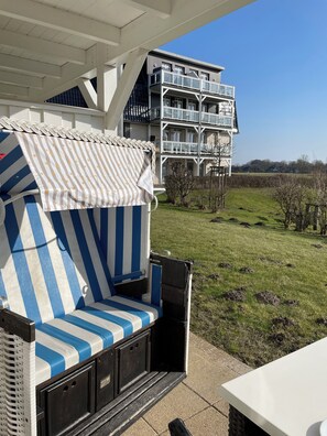 A covered outdoor seating area with a striped sofa and a table, overlooking a grassy field and a multi-story building. - "Moin im Deichgraf" Apartment on the Beach and at the Harbor in Wremer (Wurster Nordseeküste)
