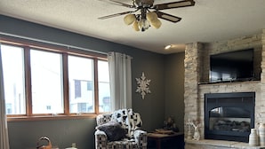 A living room with a stone fireplace, a ceiling fan, a patterned armchair, and a view of the outside through large windows. - Cozy and quaint farmhouse on an acreage  (Mountain Lake)