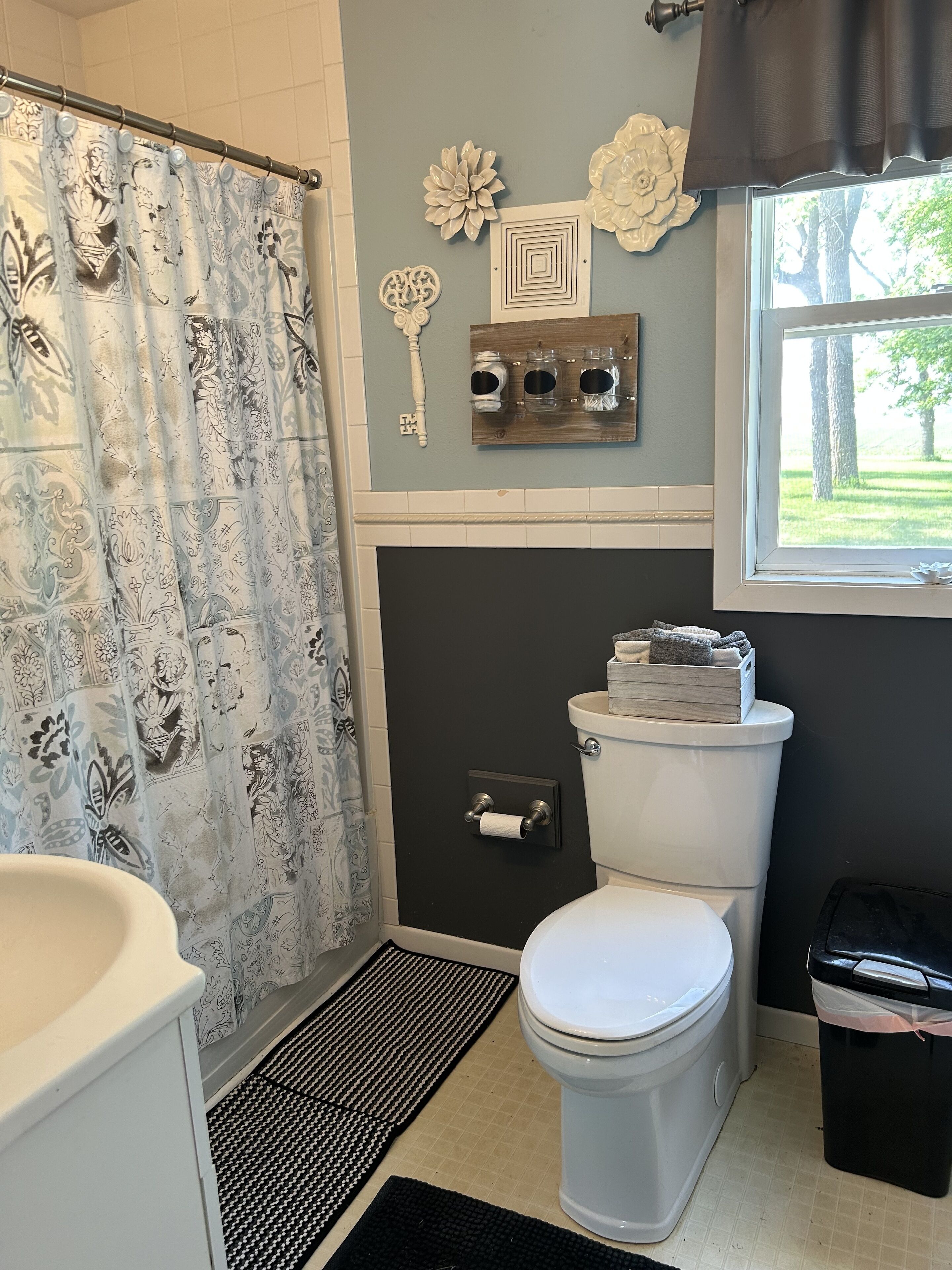 A bathroom with a toilet, a shower curtain, a sink, a window with a view of trees, and decorative wall art.