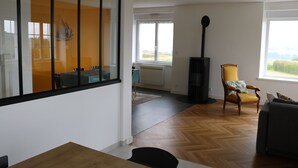 Una sala de estar moderna con piso de madera, un comedor con mesa y sillas, un sillón amarillo y un ventanal con vista a zonas verdes.