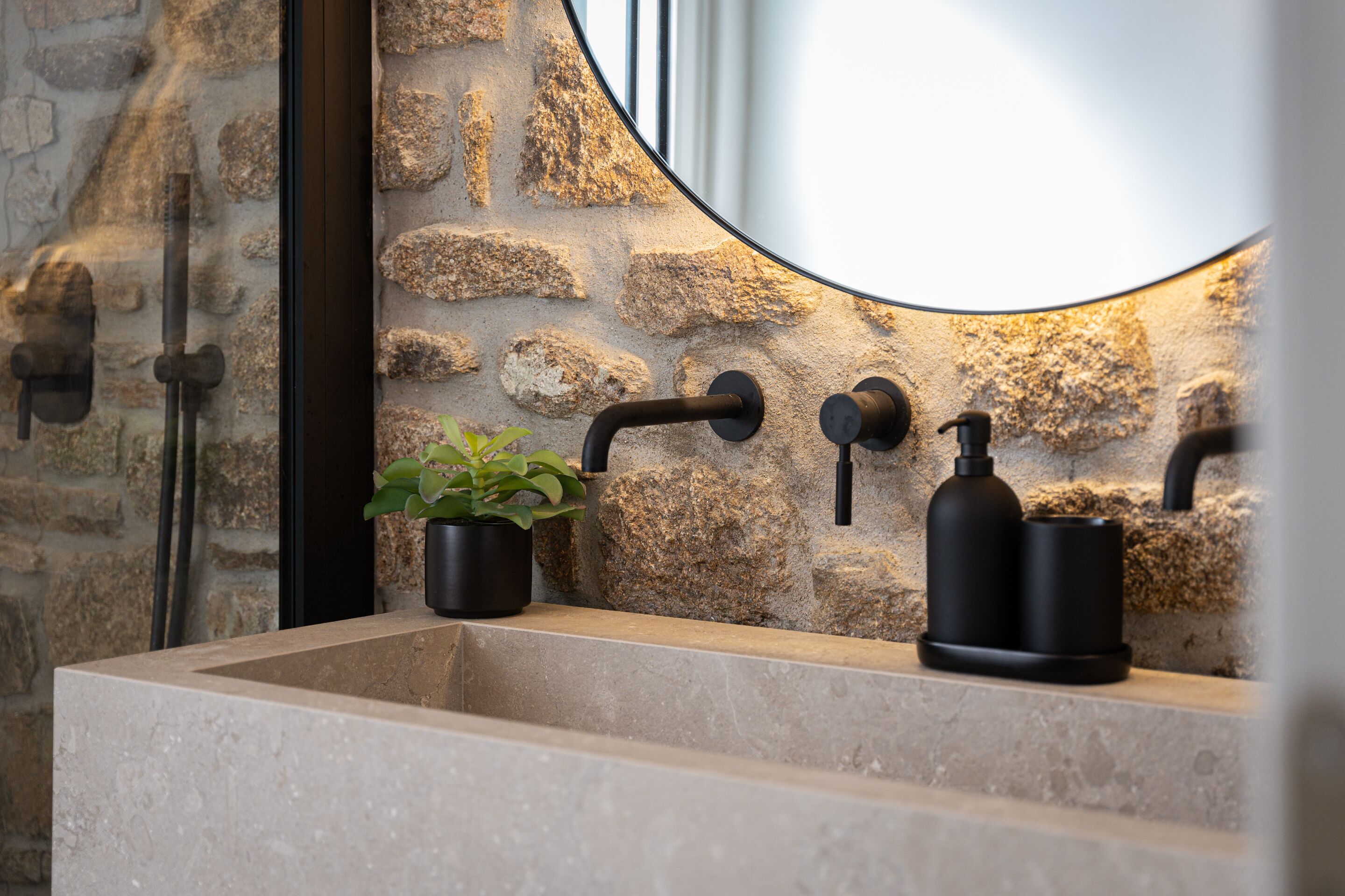 A bathroom with a stone wall, a round mirror, a sink, and a potted plant.
