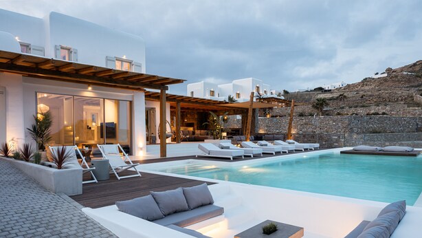 Zona de piscina exterior moderna amb gandules i una zona de seients, envoltada per una paret de pedra i una pèrgola de fusta.