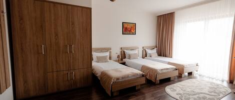 A hotel room with two single beds, a wardrobe, a large window with curtains, and a wooden floor.