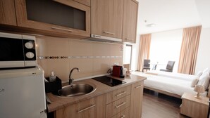 Une cuisine moderne avec des meubles en bois, un four à micro-ondes encastré et un évier. La pièce adjacente contient un lit, une table de chevet et une lampe.
