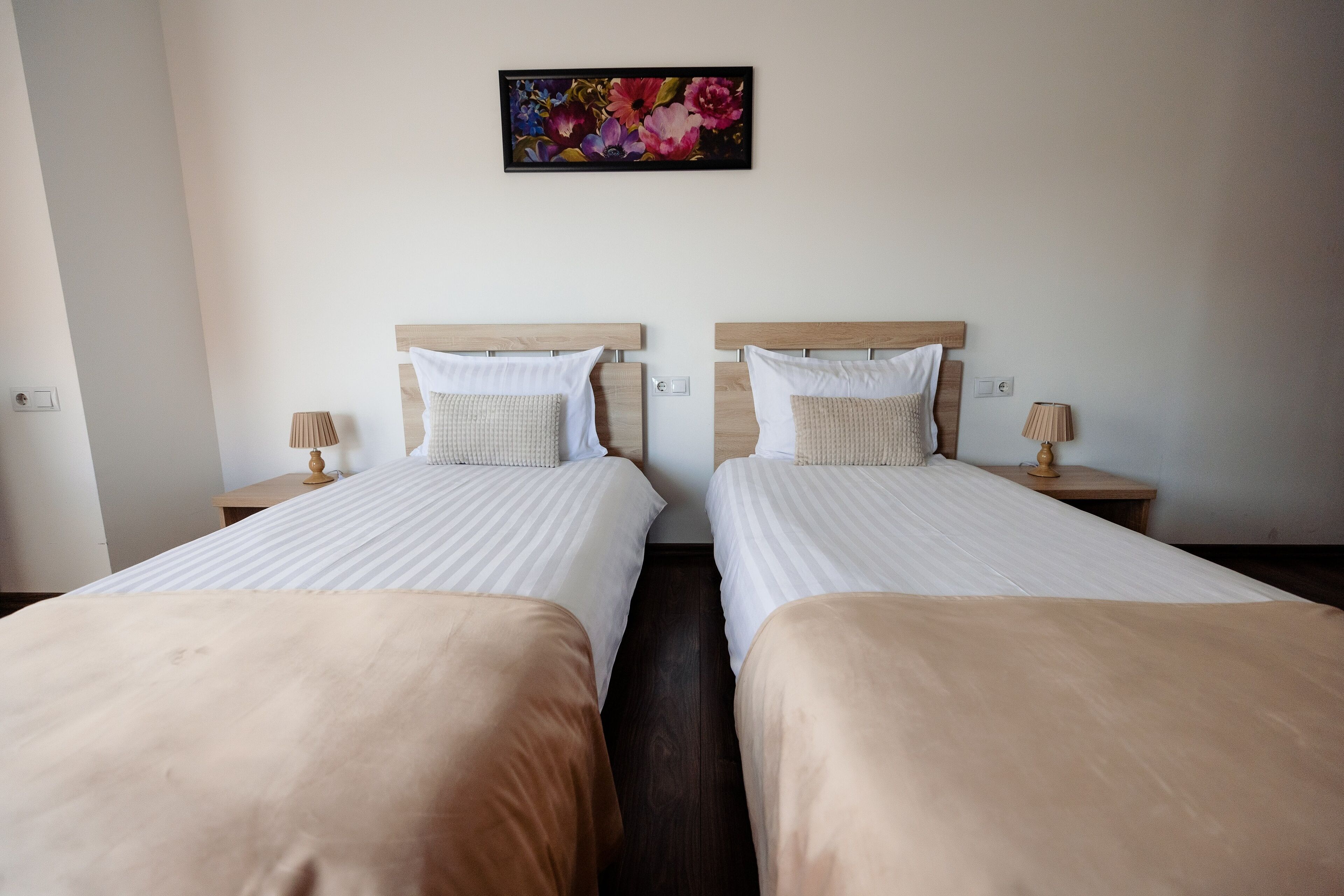 A hotel room with two beds, each with a wooden headboard, white bedding, and a beige throw blanket. There is a floral artwork on the wall above the beds, and two bedside lamps on wooden nightstands.