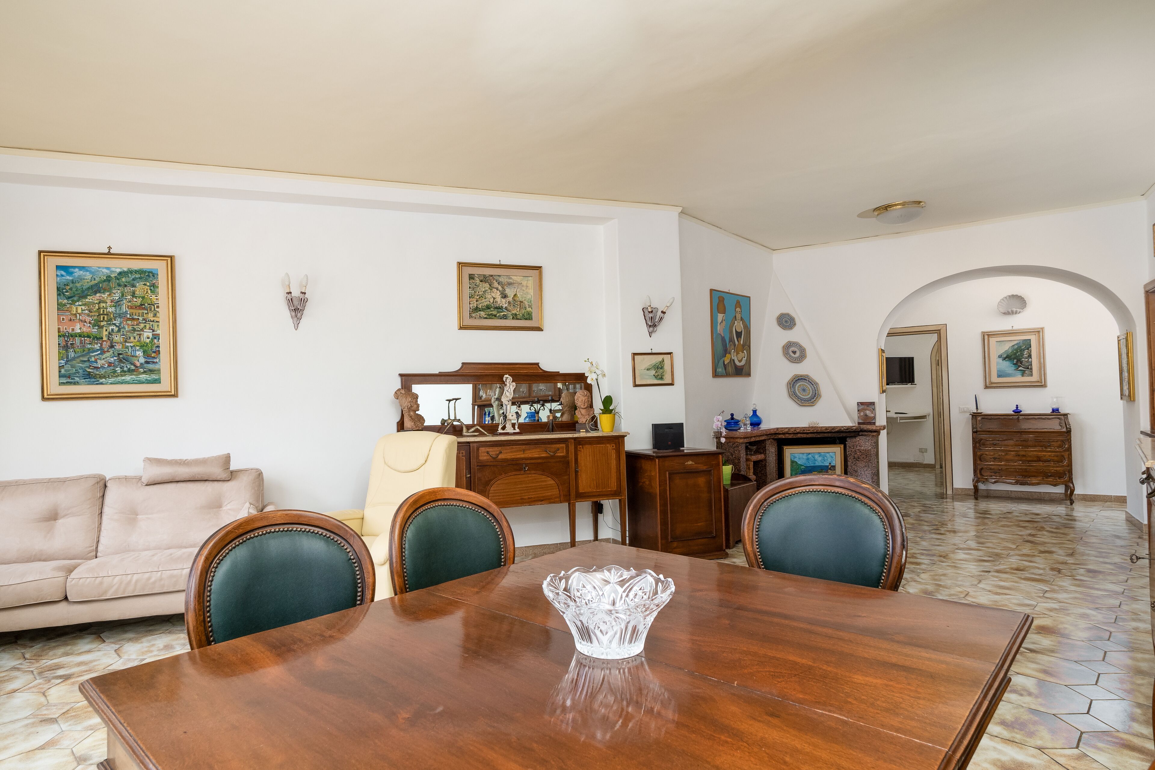 A dining area with a wooden table, chairs, and a vase. There is a sofa and a cabinet with various items on top.