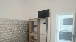 A room with a stone wall, a ceiling fan, a TV on a wooden stand, and a kitchen visible through an open door.