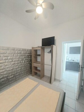 A room with a stone wall, a ceiling fan, a TV on a wooden stand, and a kitchen visible through an open door.