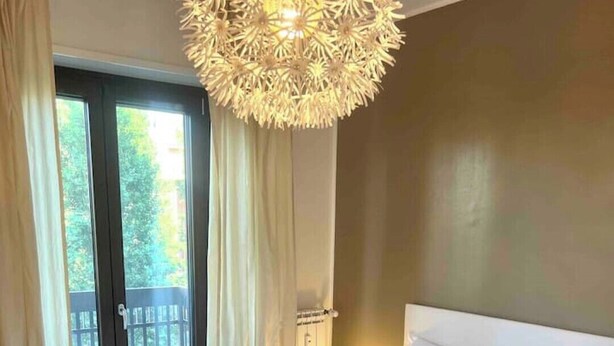 Et hotelværelse med en stor seng, hvide sengetøj, en dekorativ loftlampe og udsigt til det fri gennem en skydedør.