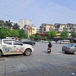 A street scene with a white van, a motorbike, and various buildings including a tall residential building and a commercial area with shops. - Blue Homestay Cau Giay (Hanoi)