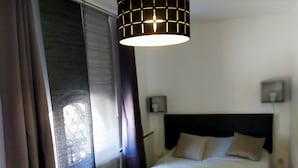 A bedroom with a bed, a hanging light fixture, a window with curtains, and a wall-mounted light. - Logement de 50 m2 en rez de Jardin (Fréjus)