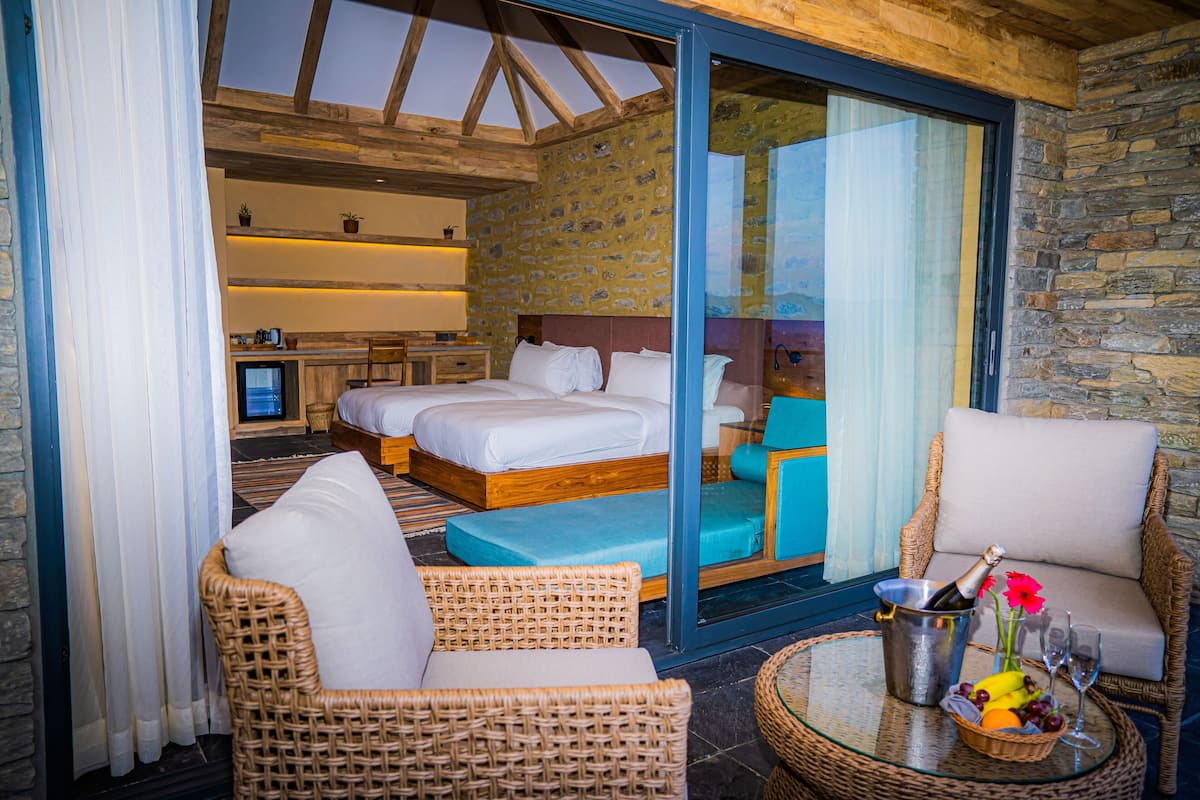 A hotel room with a large bed, two bedside tables, a television, a wicker chair, a glass table with a fruit basket and champagne, and a stone wall.