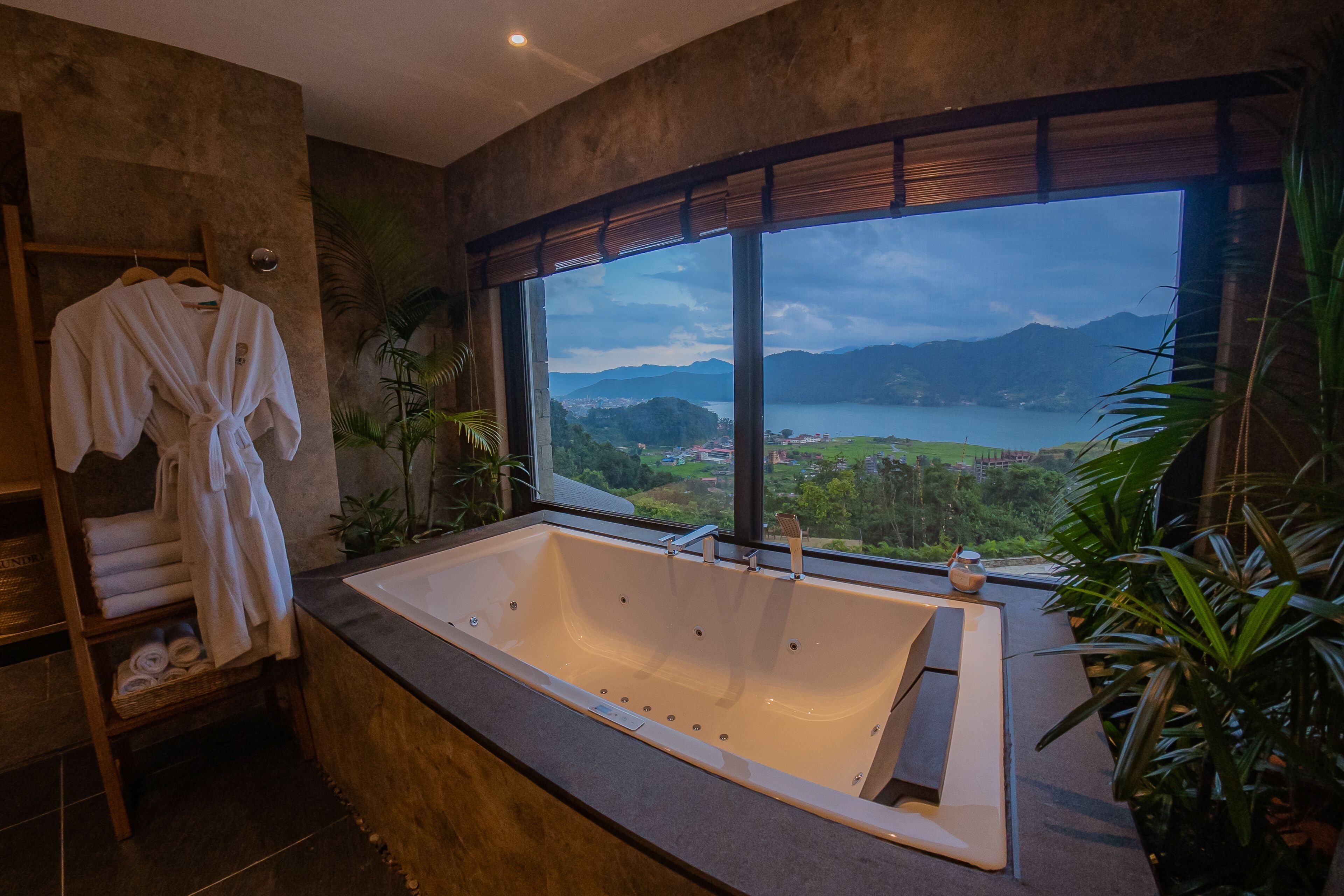 A bathroom with a large bathtub, a window with a view of a lake and mountains, and a shelf with towels.