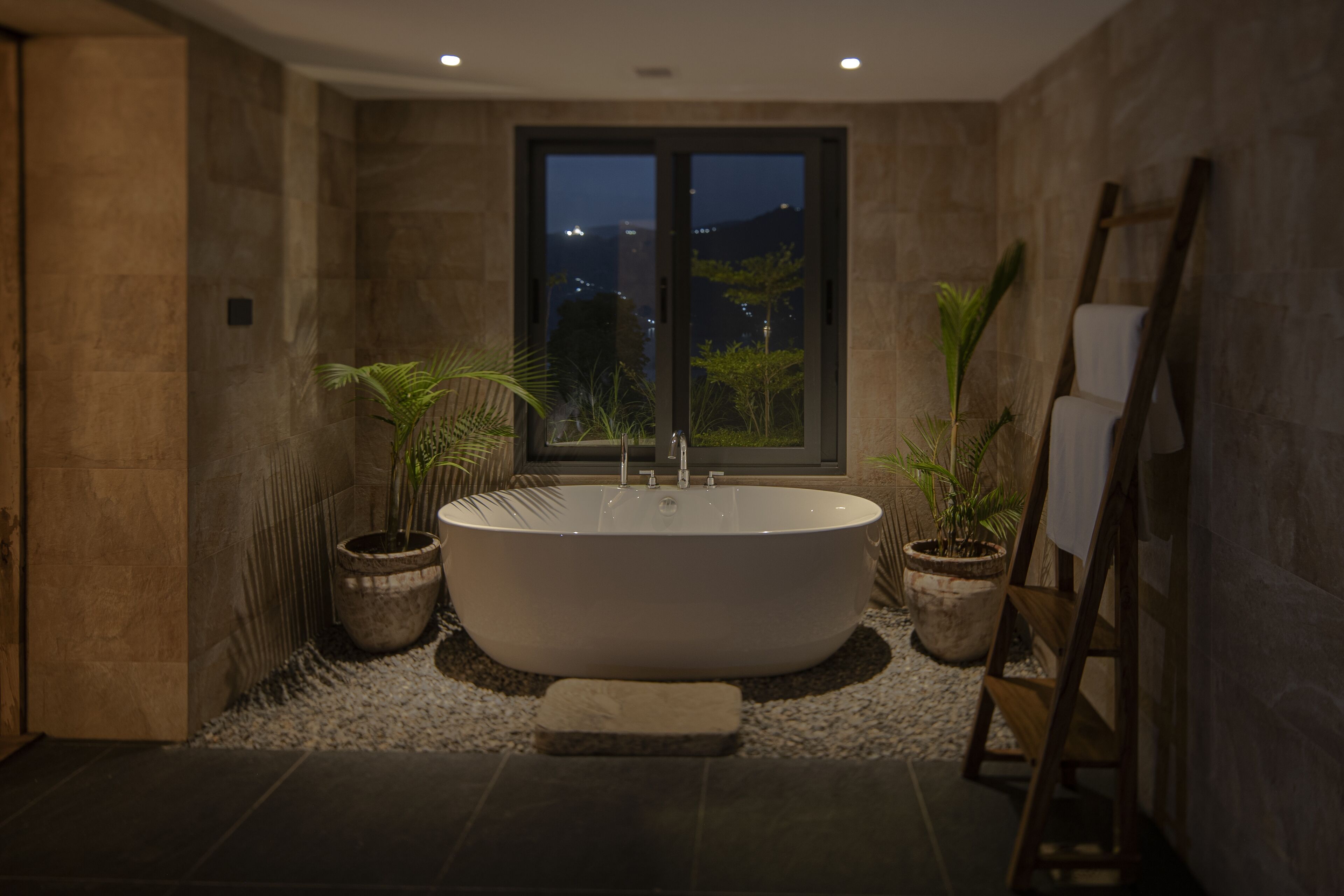 A modern bathroom with a freestanding bathtub, two potted plants, a wooden ladder, and a view of the city at night.