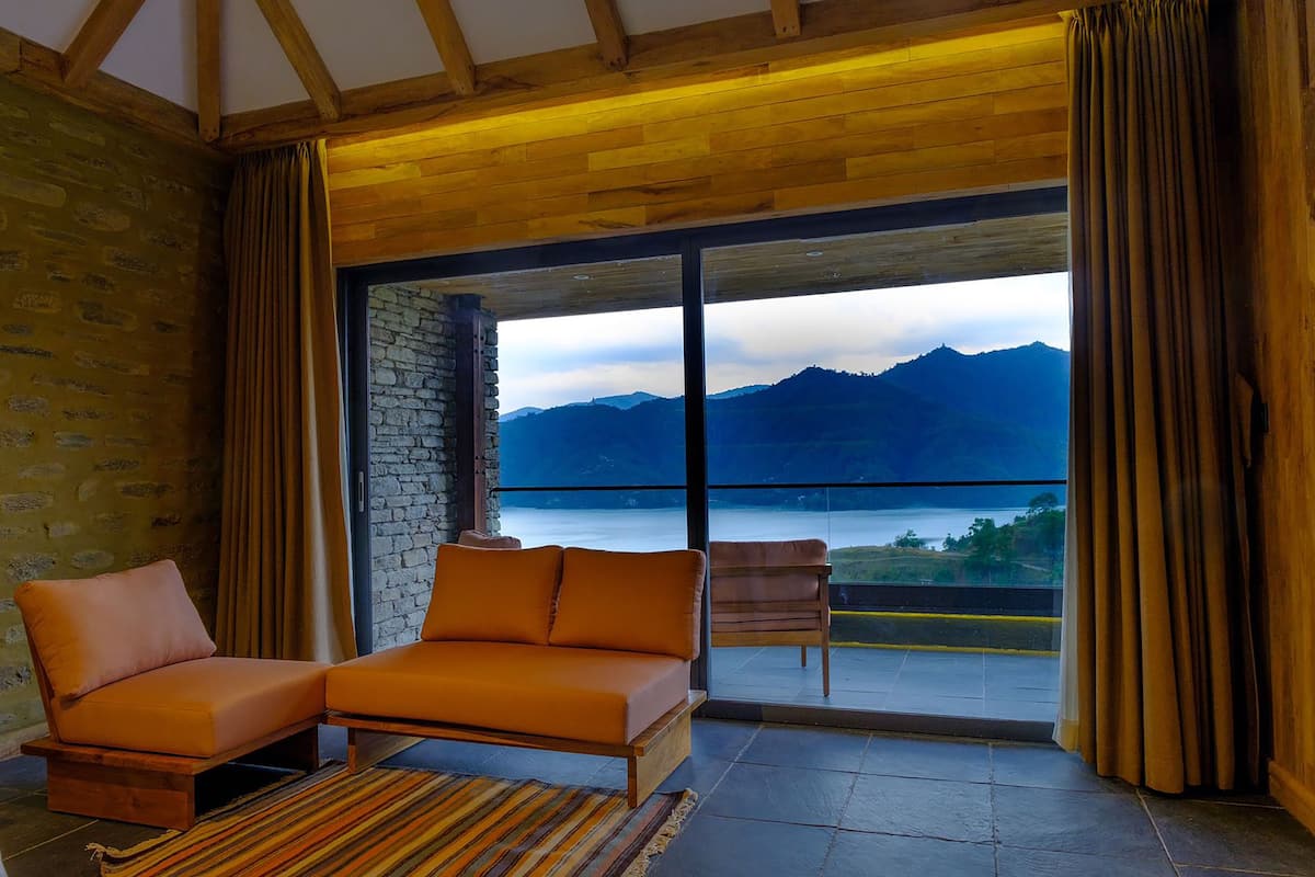 A room with a wooden ceiling, a stone wall, a sofa, a chair, and a large sliding glass door leading to a balcony with a mountain view.