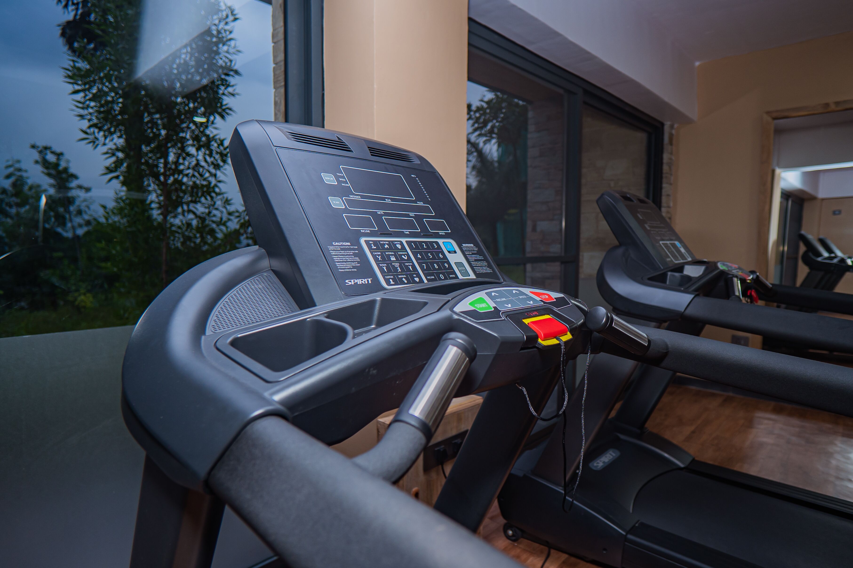A treadmill with a digital display and control panel, positioned in a room with a large window and a view of trees outside.