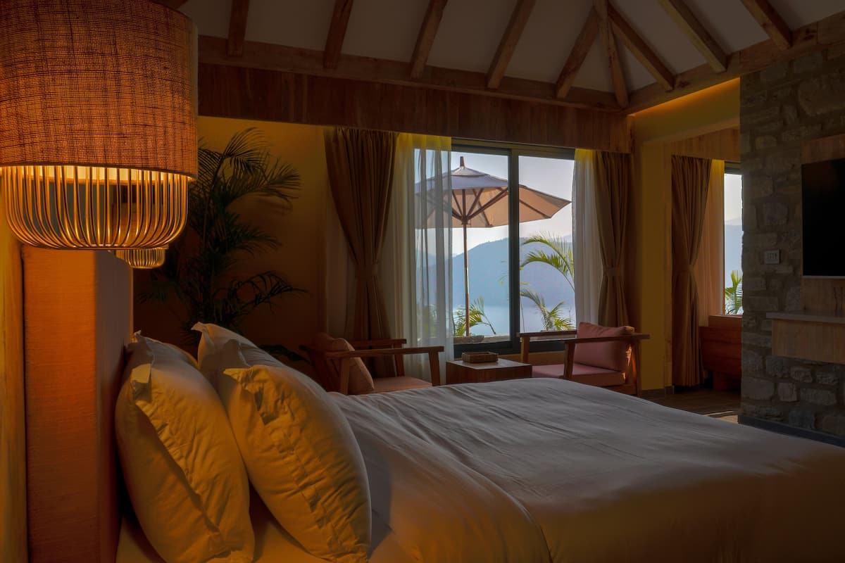 A bedroom with a large bed, a TV mounted on a stone wall, and a view of the sea through a window.