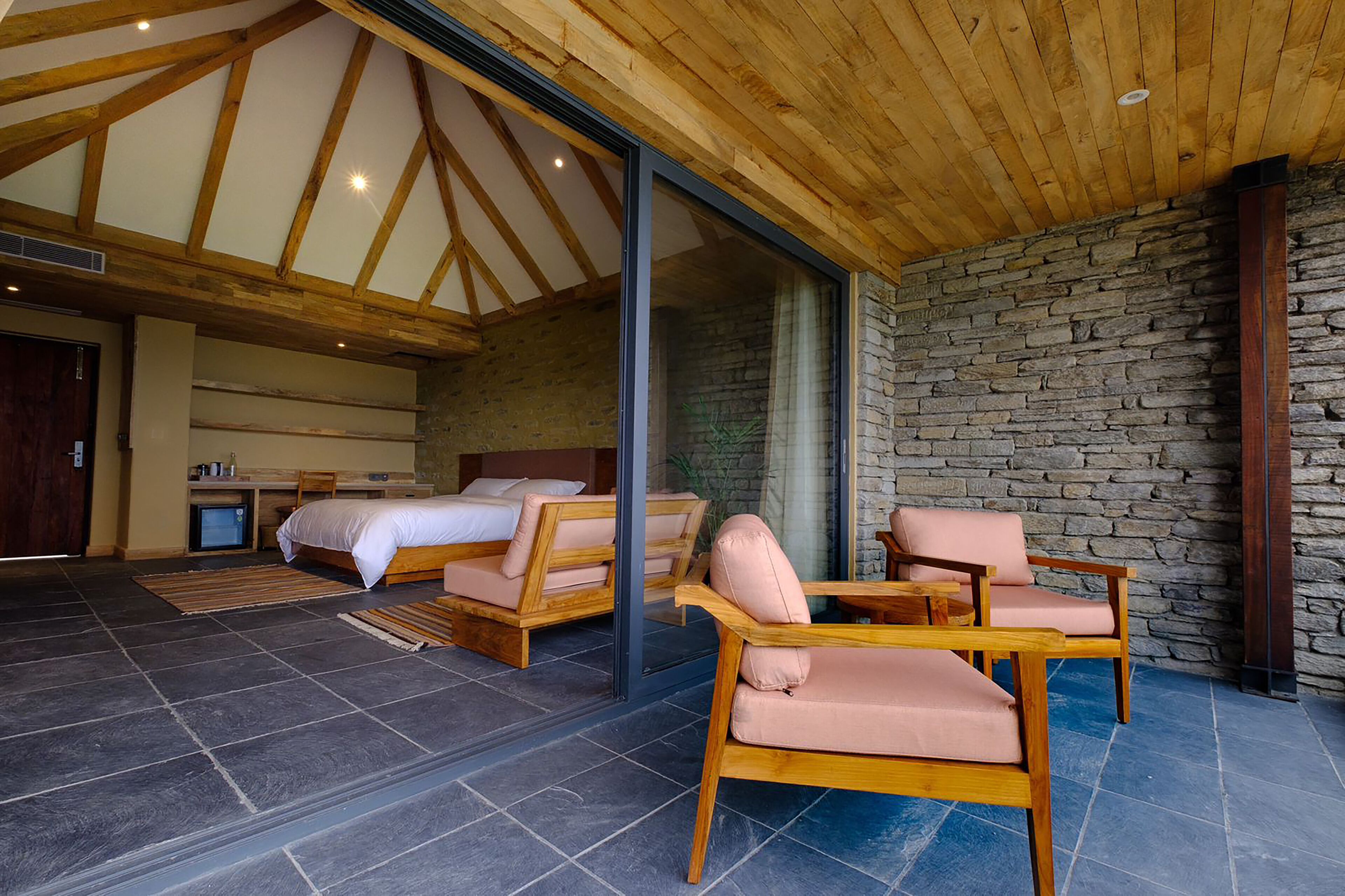 A spacious room with a high ceiling, stone walls, and a large window. There are two beds and a seating area with chairs.