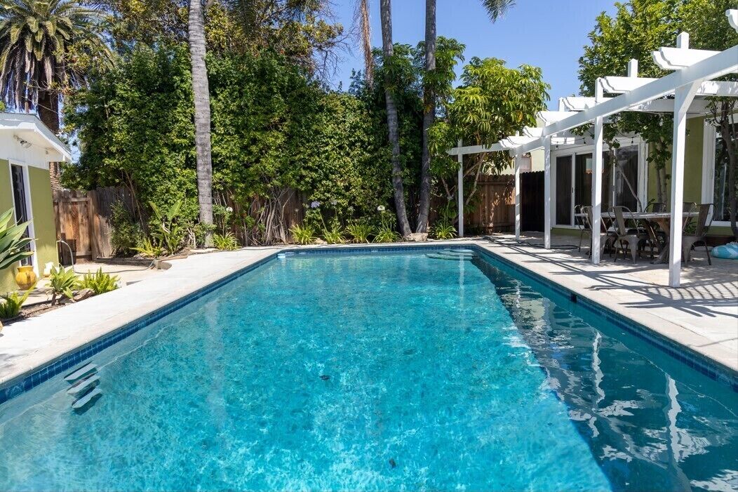 A rectangular swimming pool with clear blue water, surrounded by a white pergola and lush greenery.