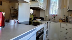 A kitchen with white cabinets, a microwave, a kettle, a washing machine, and a window with a curtain.