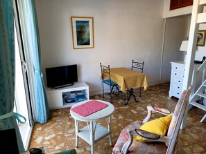 Quarto mobiliado com televisão, mesa e cadeiras, um pequeno armário branco e uma poltrona estampada.