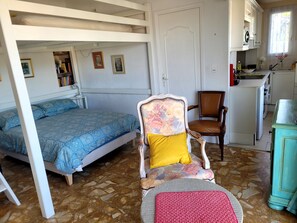 Quarto com cama, uma cadeira com almofada floral, uma mesinha e uma pequena cozinha.