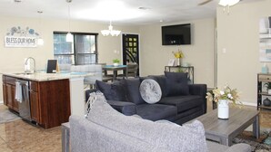 Una sala moderna con un sofá seccional gris, una mesa de centro y un televisor montado en la pared. Cuenta con una zona de cocina con una barra y una mesa de comedor.