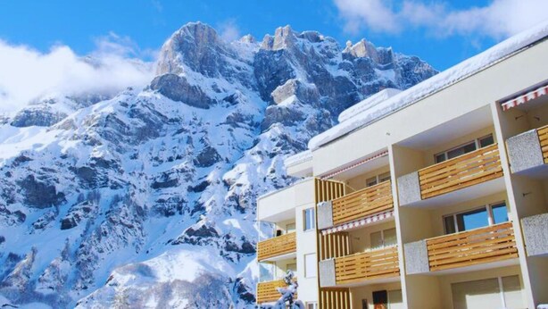 A hotel with a mountain backdrop, featuring balconies with wooden railings and a clear blue sky. - Haus Cristal  (Leukerbad)