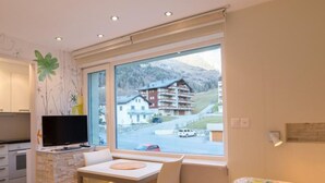 A modern room with a dining area, a bed with floral bedding, a TV, and a view of a mountain and buildings. - Haus Cristal  (Leukerbad)